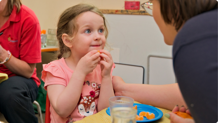 Healthy Eating in the Early Years - a 3 year old girl being encouraged to eat the orange peeled for her by a Poppies Daycare Nursery practitioner 