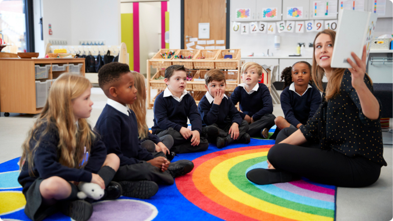 How to apply for primary school: Female teacher holding up a book in front of her class of elementary school kids sitting on the floor in a classroom, side view