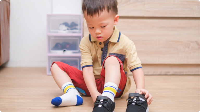 Activities to Promote Independence in Toddlers shown by todler sat on a wooden floor putting on their shoes on their own