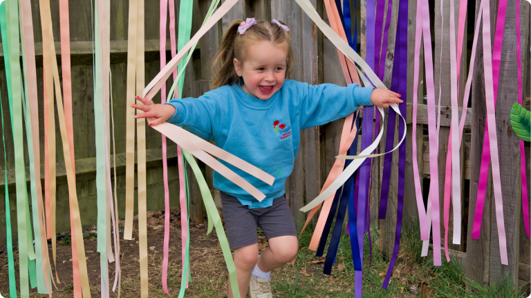 How to Build Confidence in a Sensitive Child shown by a little girl running through streamers in the garden at our Perham Nursery 