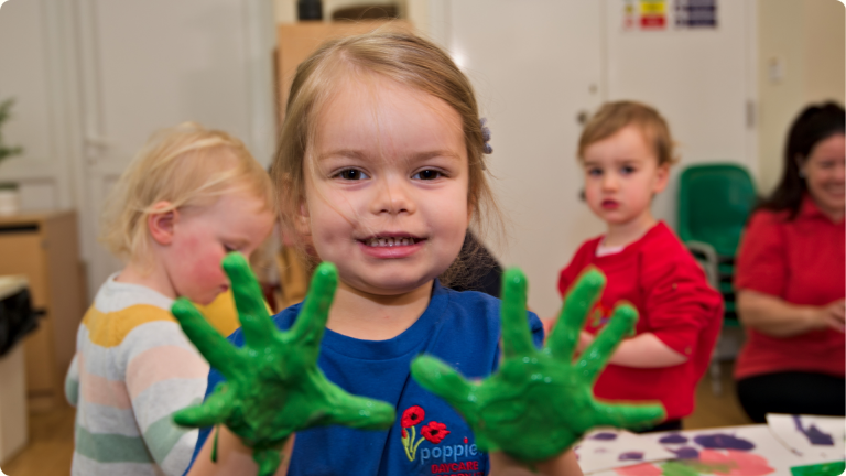 How to Build Confidence in Children shown by a little girl with green paint on her hands and showing them to the camera with glee